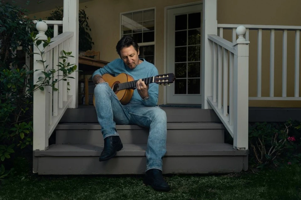 Adult male playing a classical guitar while sitting on the steps of a house with a lawn and plants. Light blue shirt and jeans.
